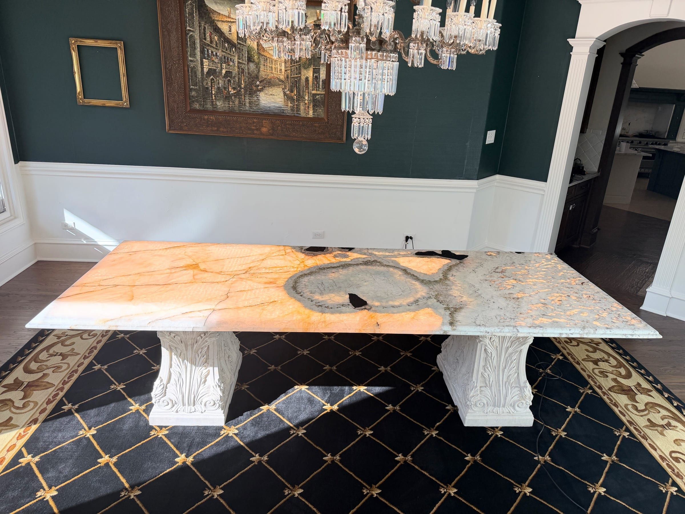A large custom dining table featuring a backlit mixed stone top with warm orange and grey tones, supported by ornate white pedestals in a formal dining room.