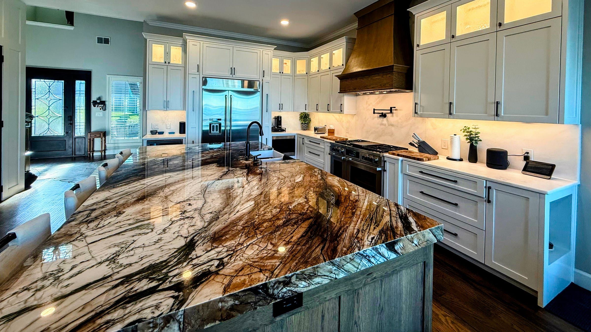 A large kitchen island featuring a dramatic brown and white granite countertop with bold veining, paired with white perimeter cabinets and a wood range hood.