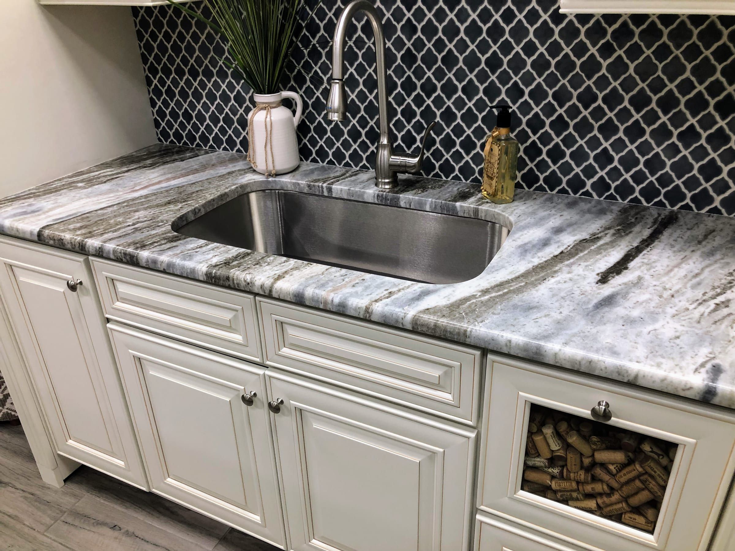 A white-cabinet wet bar topped with grey and brown Fantasy Brown marble, an undermount stainless sink, and a black arabesque tile backsplash.