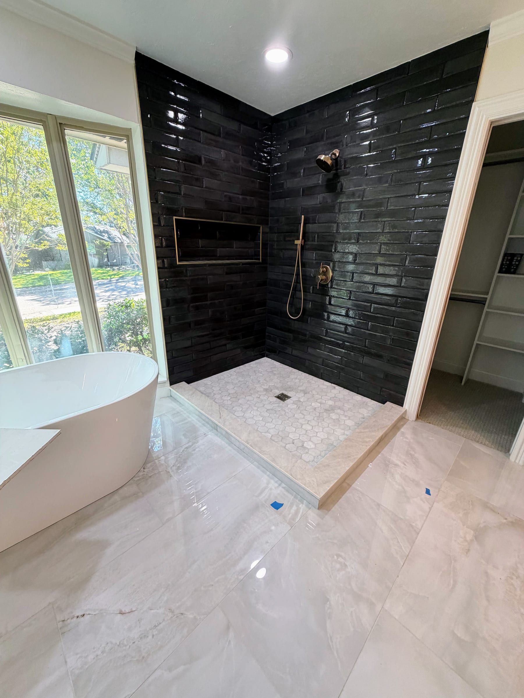 Modern bathroom featuring light marble floor tiles, a white soaking tub, and a shower enclosure with a marble mosaic tile pan and glossy black subway tile walls.