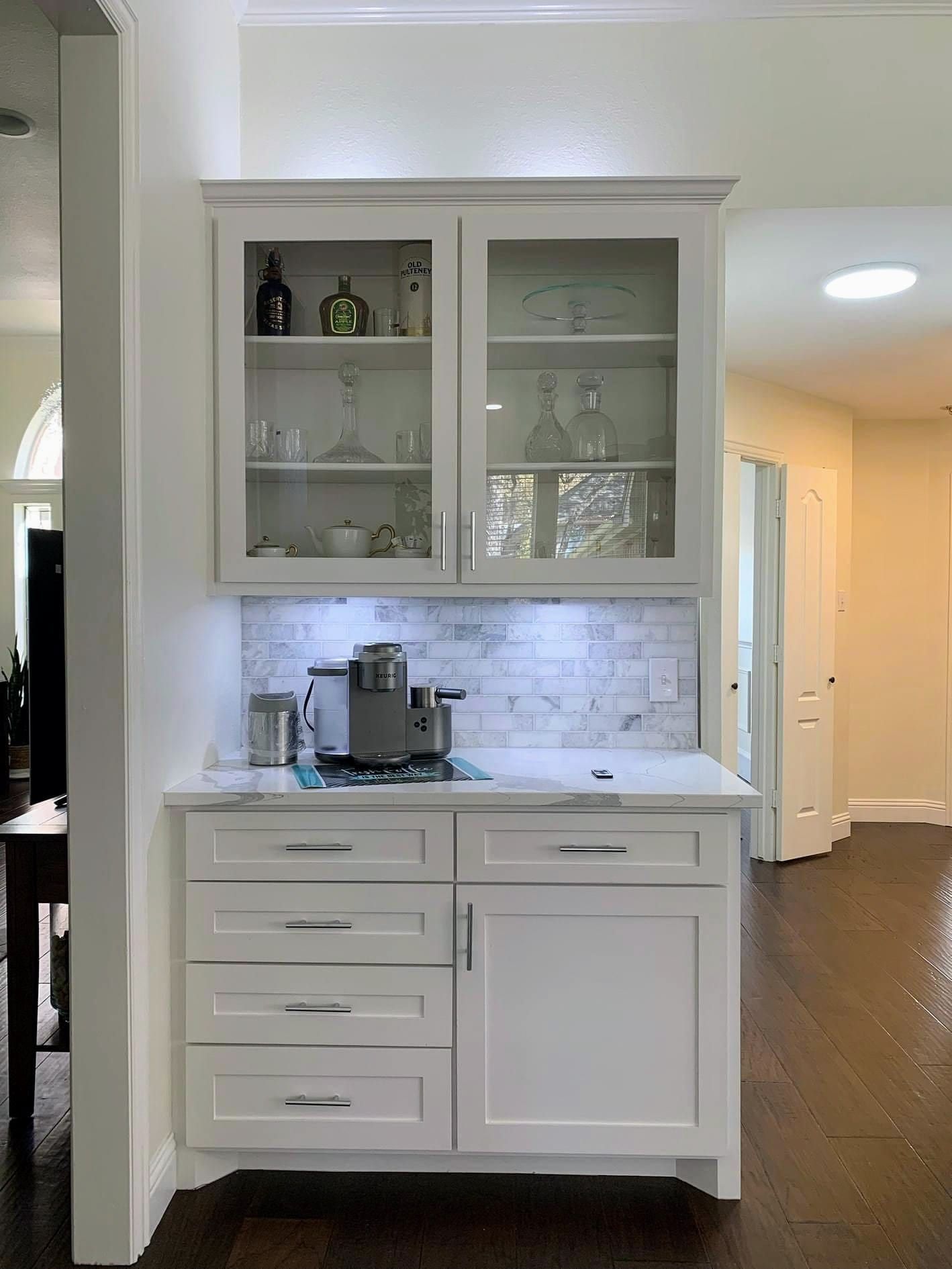 A white quartz countertop with subtle grey veining installed in a kitchen coffee bar area with white cabinets and a marble tile backsplash.