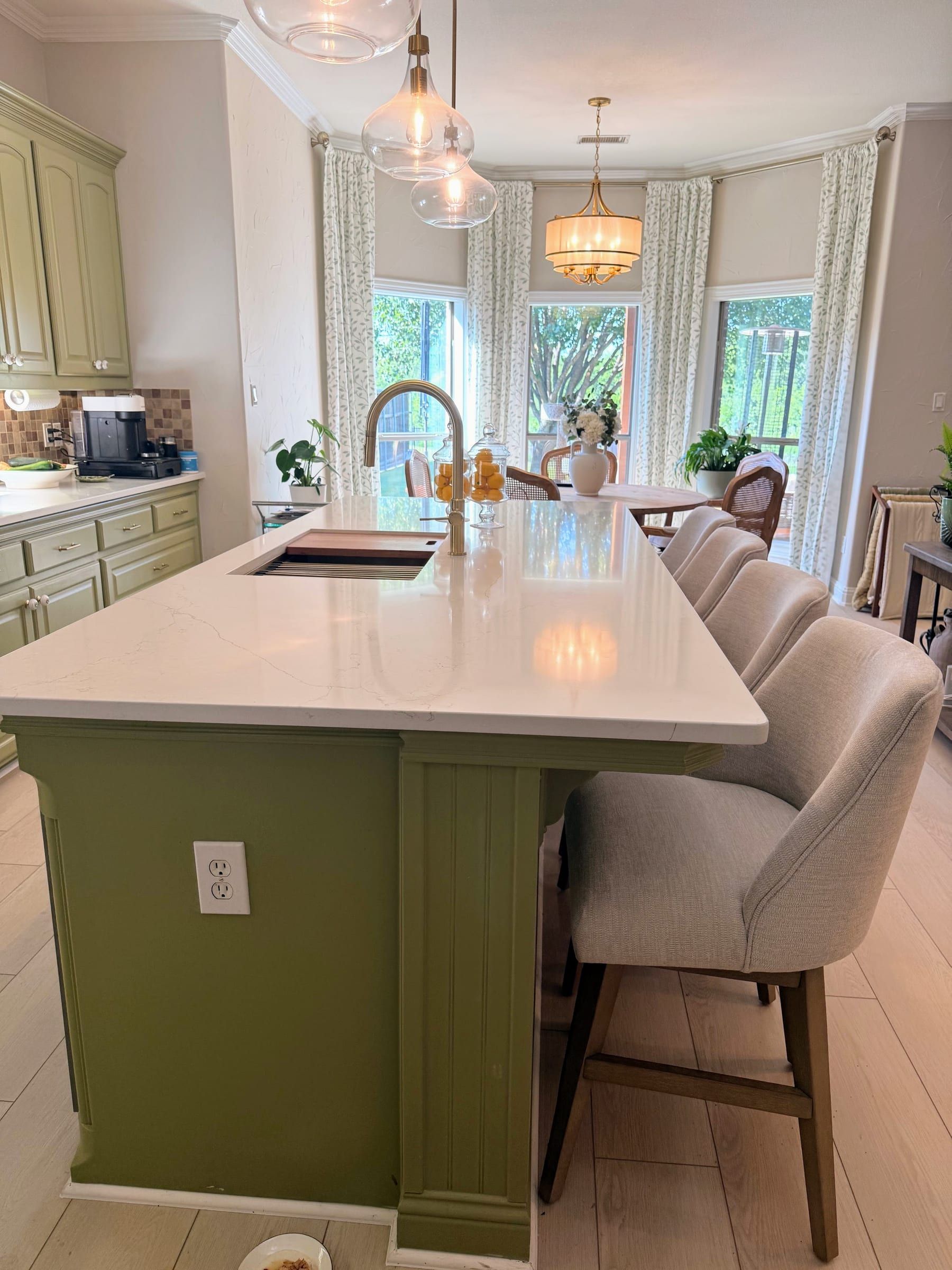 A white quartz kitchen island with subtle grey veining and a gold faucet, paired with sage green cabinetry and upholstered bar stools.