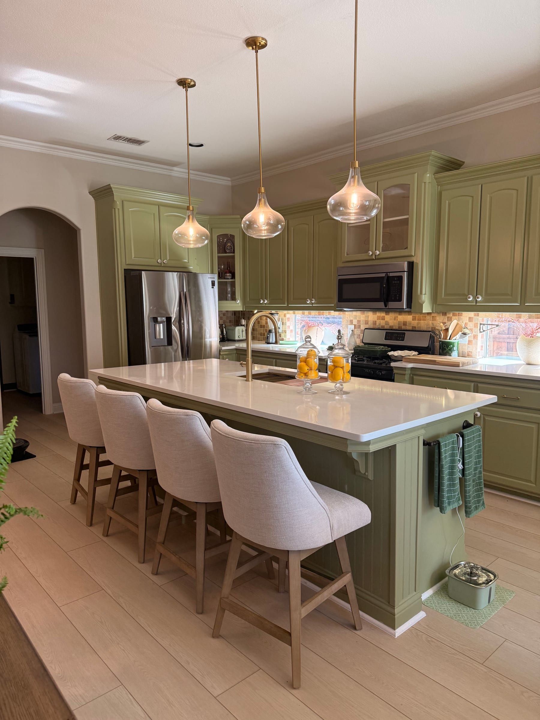 A large kitchen island with a white quartz countertop and gold faucet, paired with sage green cabinetry and light wood flooring.