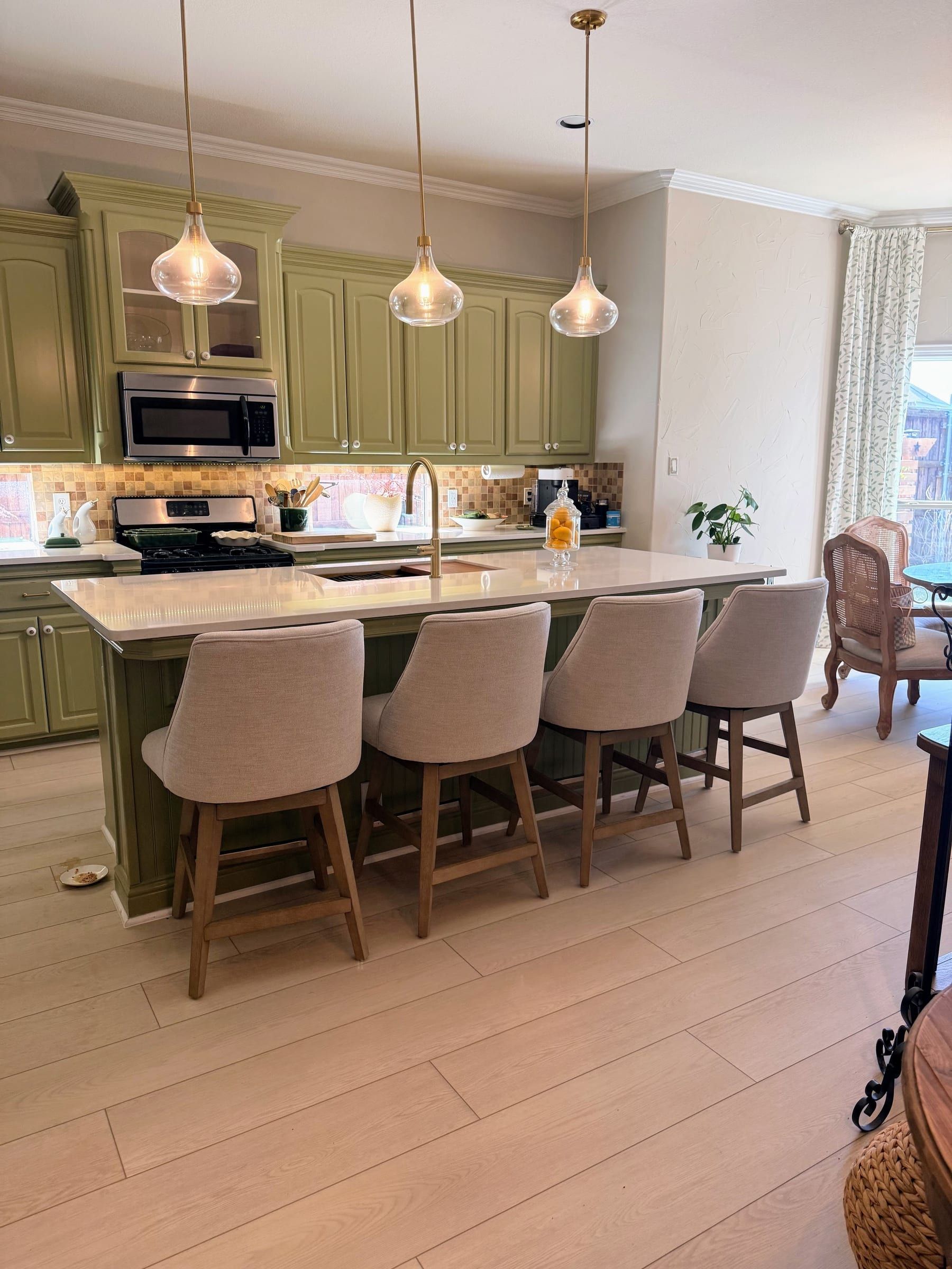 A large white quartz kitchen island with a gold faucet sits in front of olive green cabinets and light wood flooring.